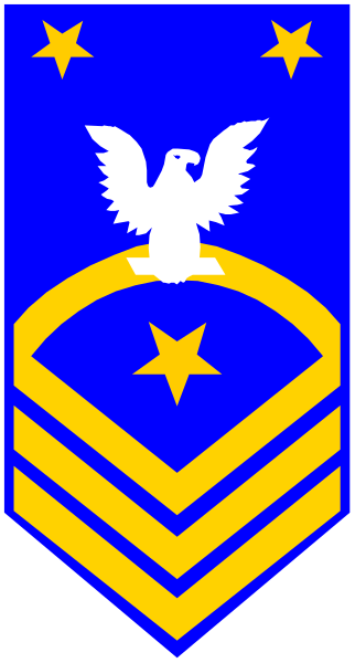 The image shows a flag with blue background featuring an eagle on top of what appears to be a shield-like emblem in gold coloring; below this is another symbol that resembles a crest, also rendered in yellow and white hues.