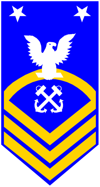 The image shows a blue flag with an emblem on it that includes what appears to be a shield bearing crossed anchors flanked by two swords; above this is another smaller shield featuring a bird in flight, and below them both there's text which seems like Latin or Greek scripture.