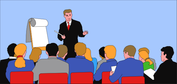 The image shows a man standing at front of an audience with his hands on hips while holding papers; he is speaking to them in what appears to be a conference room environment.