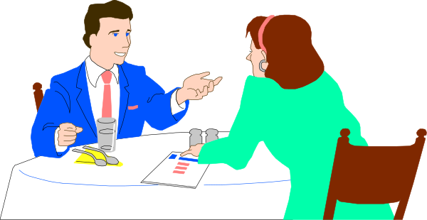 The image shows a man sitting at a dining table with his hand raised as if he's speaking to someone who is standing across from him; both individuals appear engaged in conversation.