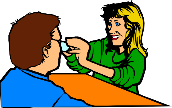 The image shows a woman with blonde hair sitting at an orange tabletop, applying what appears to be makeup on someone's face who is seated across from her in front of another person standing behind them both.