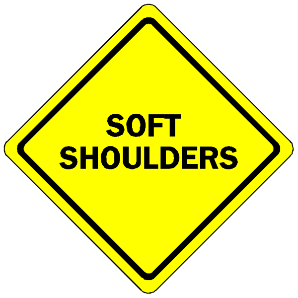 The image shows a yellow diamond sign with black text that reads "SOFT SHOULDERS" on top of another smaller square sign below it labeled in white letters as "SLOW DOWN."