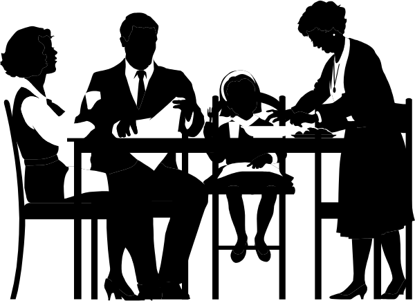 The black and white photo shows a group of four individuals sitting around an oval table with various items on it; there is also another person standing in front of them.