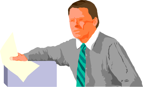 The image shows a man standing at an office desk with papers on it; he is pointing to something off-screen while looking downward towards his left hand side.