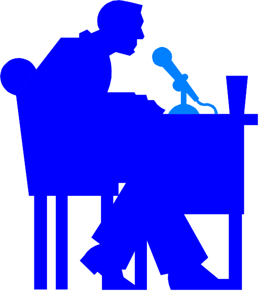 The image shows a man sitting at a desk with his hands on it while speaking into an open microphone placed before him in front of what appears to be a podium. He is wearing glasses and has short hair, dressed casually yet professionally for public speaking or interviewing purposes.